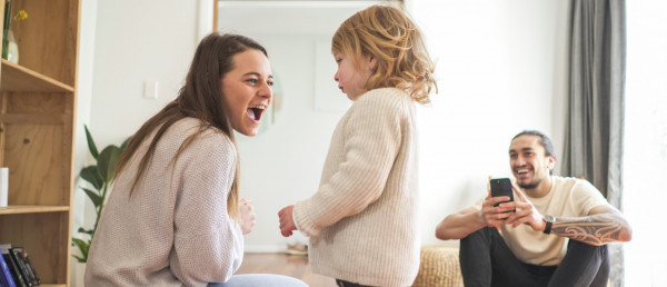 A mother and daughter playing at home with the father taking a picture on his phone