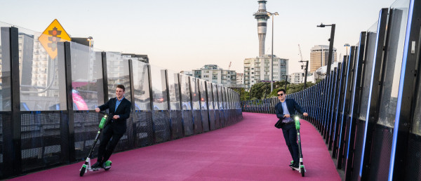 Two men on scooters on a bridge, with Auckland in the background