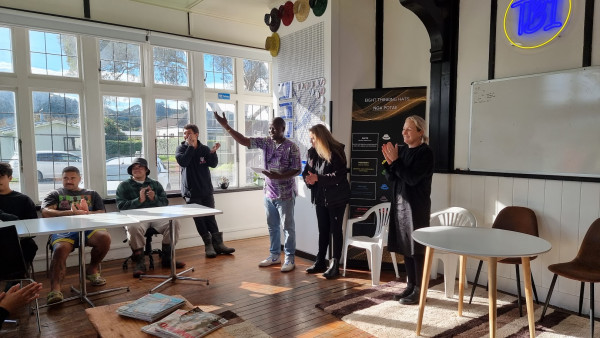 Six people in a sun-filled room, three seated by a window at table, three standing, everyone clapping